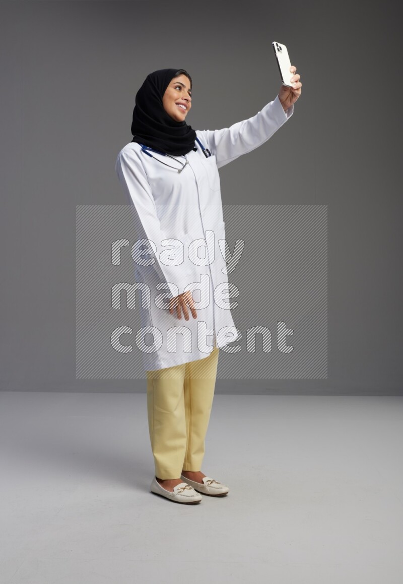 Saudi woman wearing lab coat with stethoscope standing taking selfie on Gray background