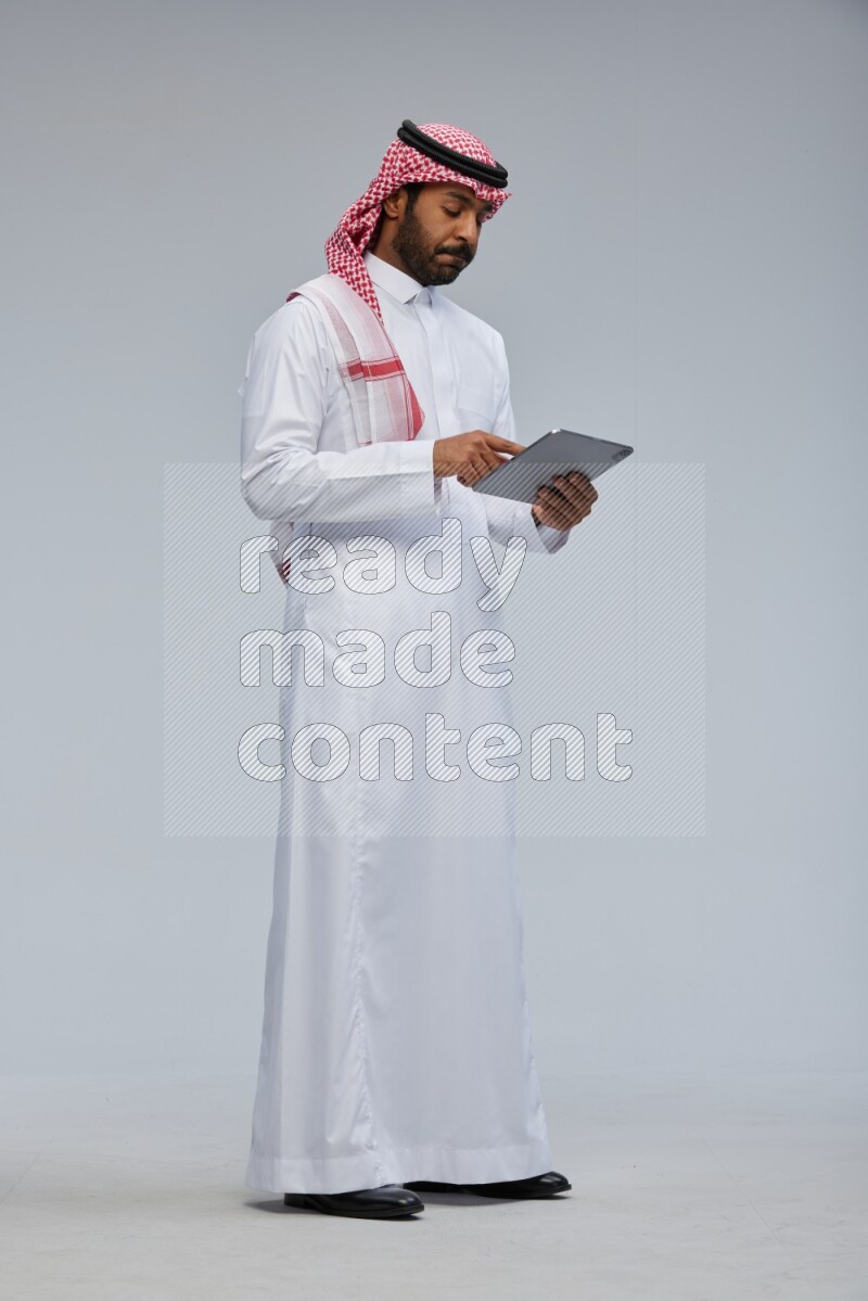 Saudi man Wearing Thob and shomag standing working on tablet on Gray background