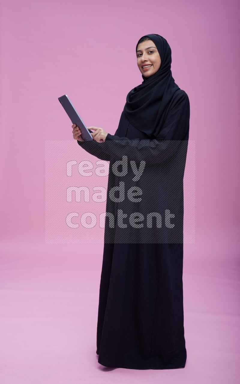Saudi woman wearing Abaya standing working on tablet on pink background