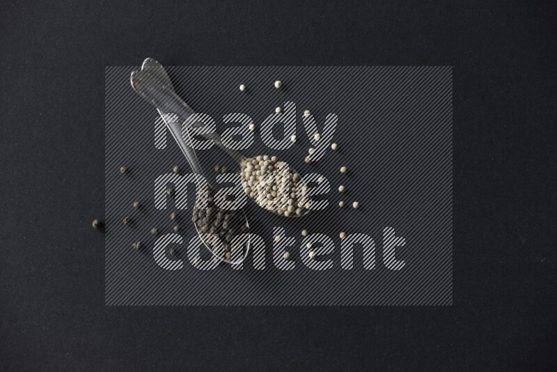 ملعقتان معدنيتان ممتلئتان بحبوب الفلفل الأسود والأبيض مع حبوب متناثرة على أرضية سوداء