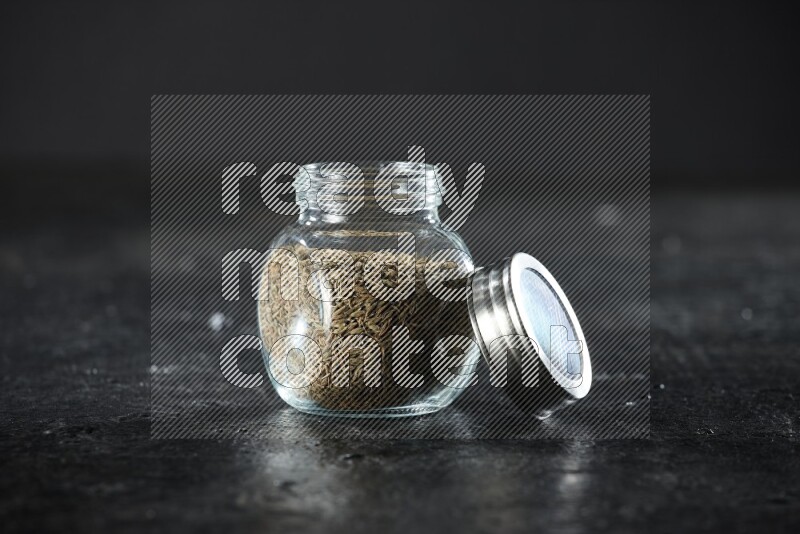A glass spice jar full of cumin seeds on a textured black flooring