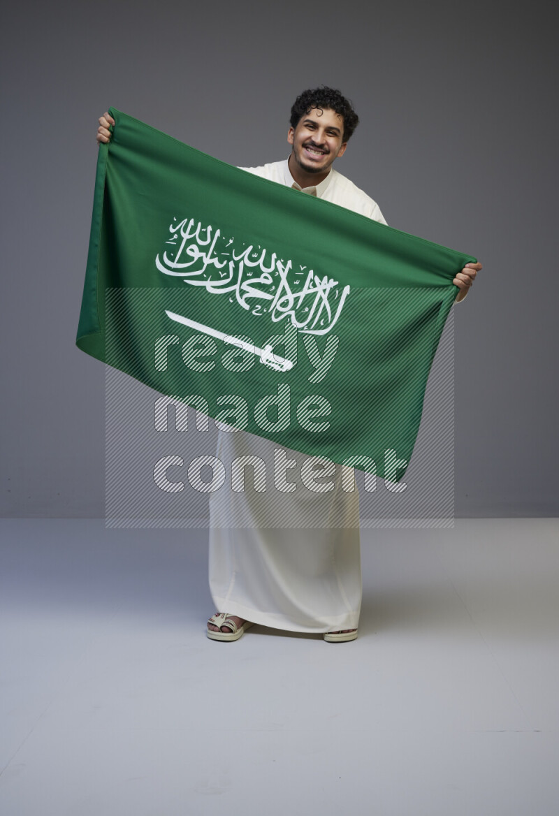 A saudi man standing wearing thob holding big saudi flag on gray background