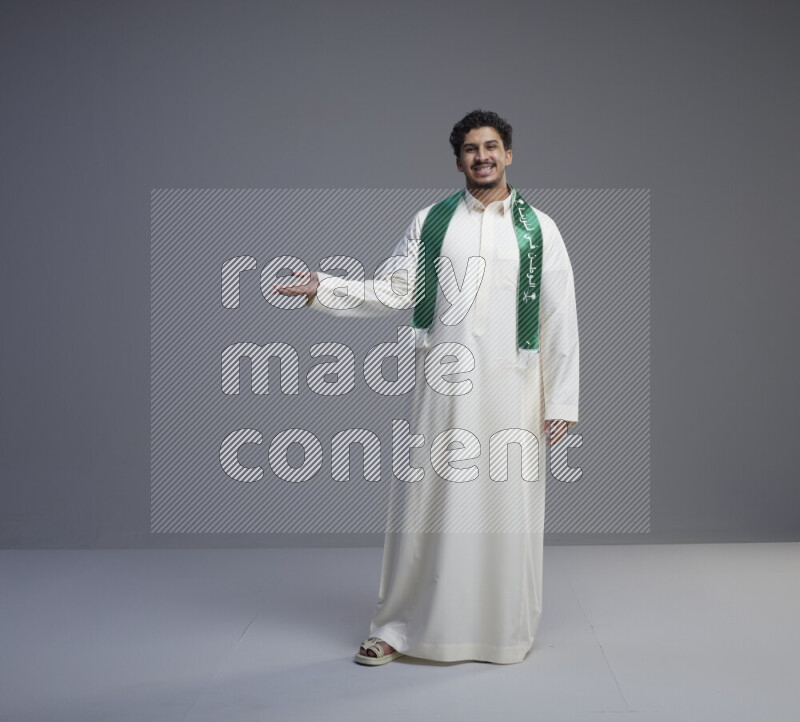 A Saudi man standing wearing thob and Saudi flag scarf on gray background