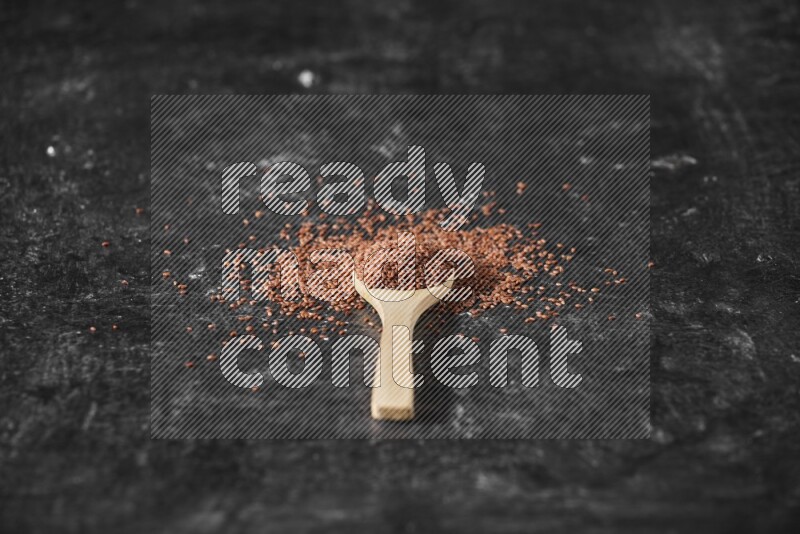 A wooden spoon full of garden cress seeds surrounded by the seeds on a textured black flooring