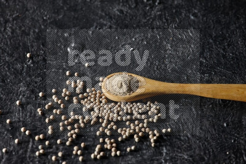 A wooden spoon full of white pepper powder with white pepper beads on textured black flooring