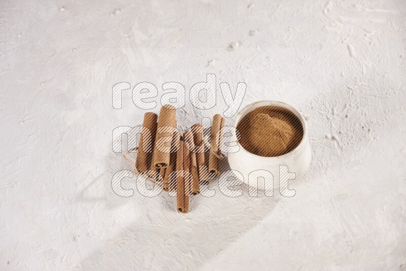Cinnamon sticks stacked beside a beige bowl full of cinnamon powder on white background