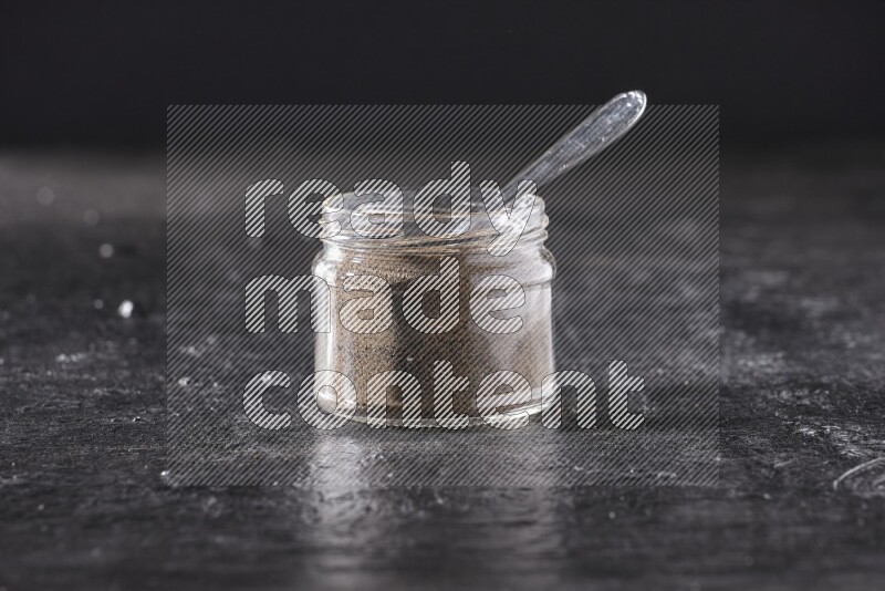 A glass jar full of black pepper powder and a metal spoon on a textured black flooring