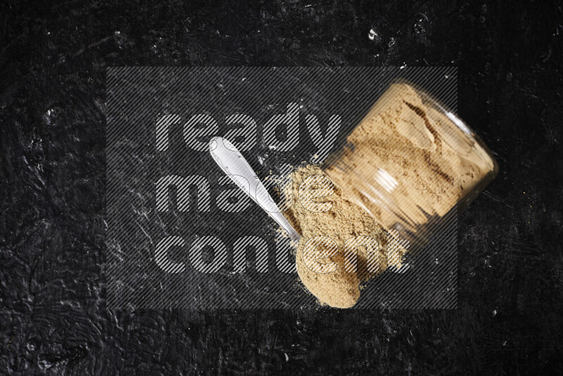 A glass jar full of ground ginger powder flipped with some spilling powder on black background