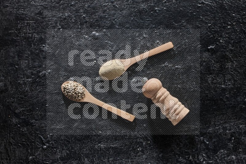 2 wooden spoons one full of white pepper powder and the other with pepper beads and a wooden pepper mill on textured black flooring