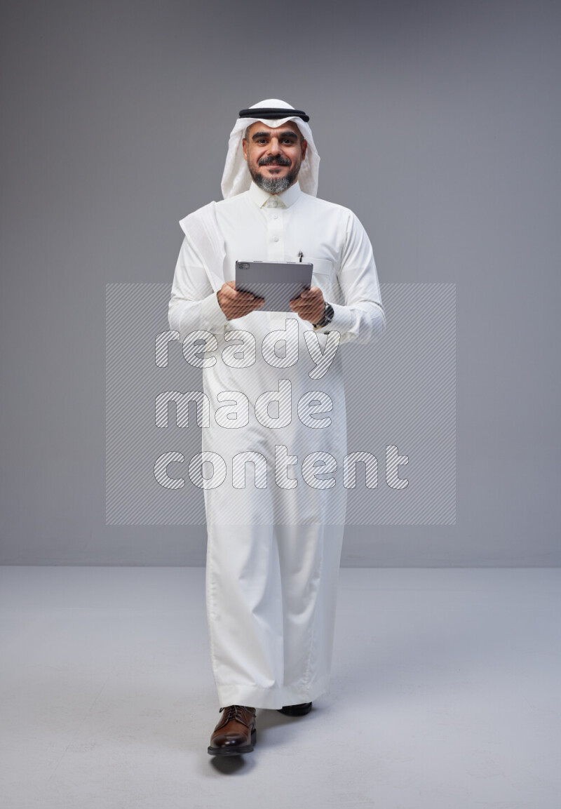 Saudi man Wearing Thob and white Shomag standing working on tablet on Gray background