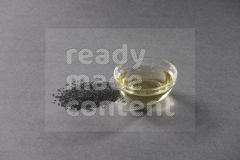 A glass bowl full of black seeds oil and black seeds beside it on a black flooring