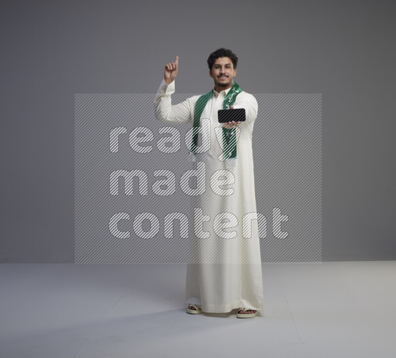 A Saudi man standing wearing thob and Saudi flag scarf showing phone to camera on gray background