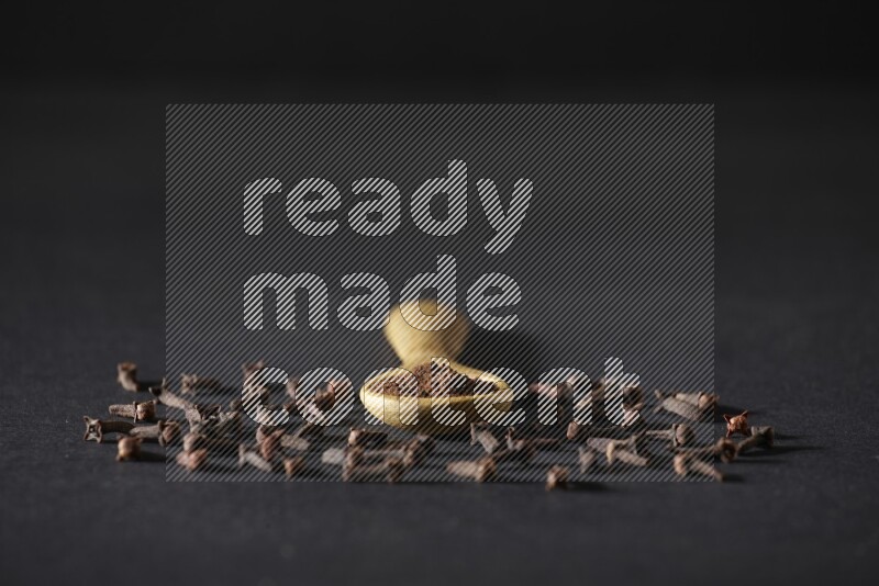 A wooden spoon full of cloves powder with spreaded whole cloves on a black flooring