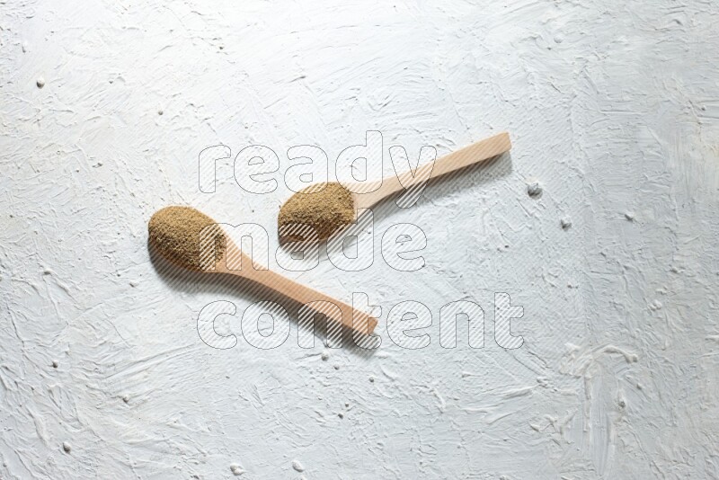 2 Wooden spoons full of cumin powder on a textured white flooring