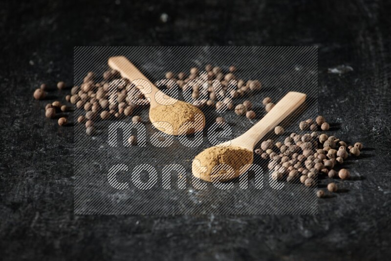 Wooden spoons full of all spice powder and allspice whole balls beside it on a textured black flooring