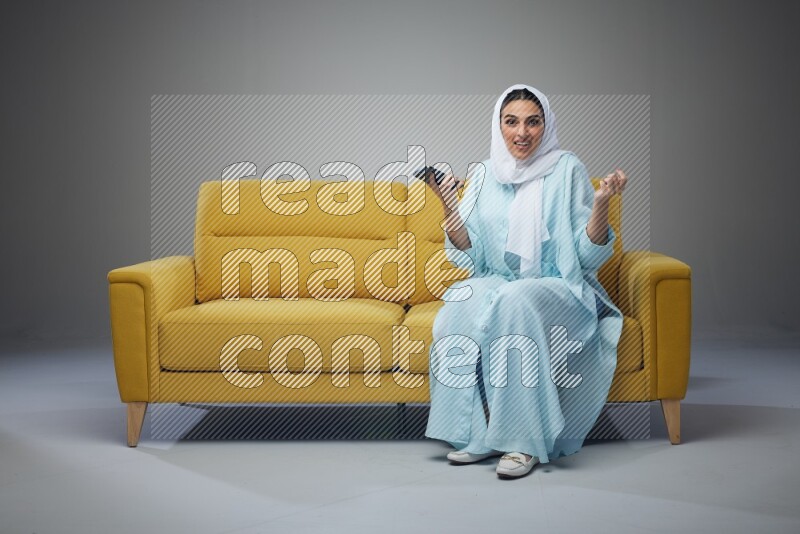 A Saudi woman wearing a light blue Abaya and white head scarf sitting on a yellow sofa and texting on her phone on a grey background