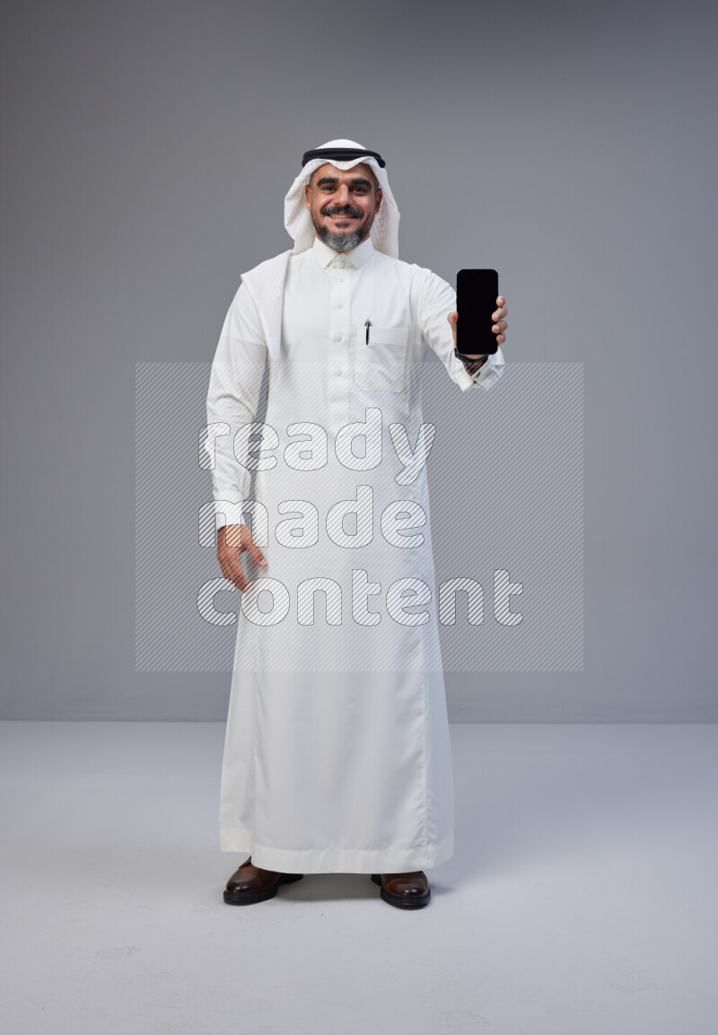 Saudi man Wearing Thob and white Shomag standing showing phone to camera on Gray background
