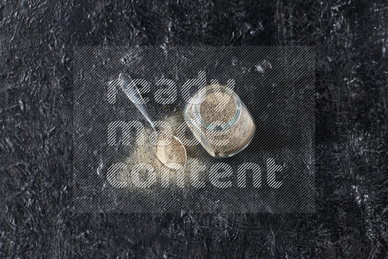 Herbal glass jar and metal spoon full of white pepper powder on textured black flooring