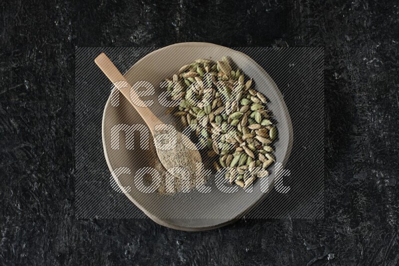 A plate filled with cardamom seeds and a wooden spoon full of cardamom powder on a textured black flooring