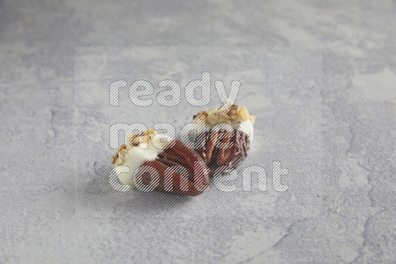 Two pecan stuffed date covered with white chocolate and chopped walnuts on alight grey background