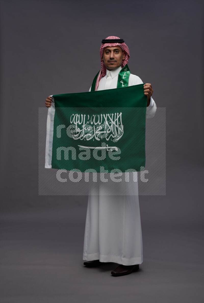 Saudi man wearing thob and shomag standing holding Saudi flag on gray background