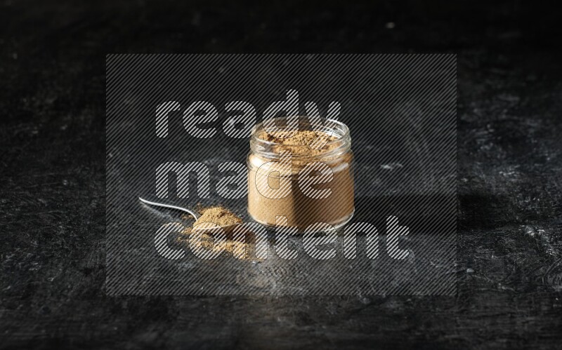 A glass jar and a metal spoon full of allspice powder on a textured black flooring