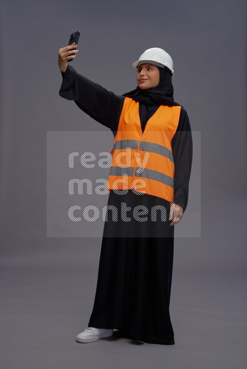 Saudi woman wearing Abaya with engineer vest standing taking selfie on gray background