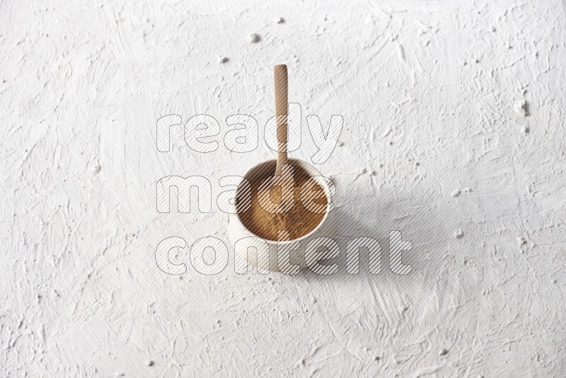 Ceramic beige bowl full of cinnamon powder with a wooden spoon on a textured white background