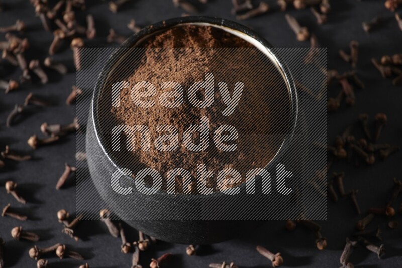 A black pottery bowl full of cloves powder and cloves whole grains on a black flooring