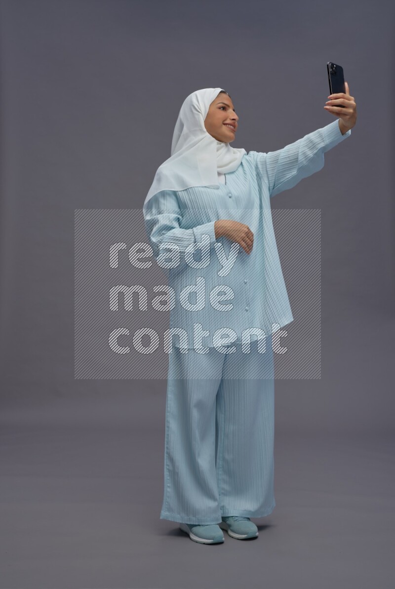 Saudi woman wearing hijab clothes standing taking selfie on gray background