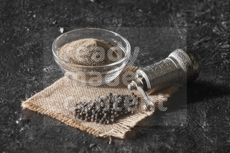 A glass bowl full of black pepper powder and black pepper beads on burlap fabric with a turkish metal pepper grinder on textured black flooring