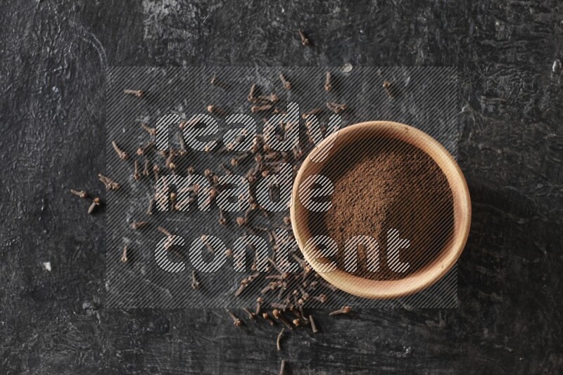 A wooden bowl full of cloves powder on a textured black flooring