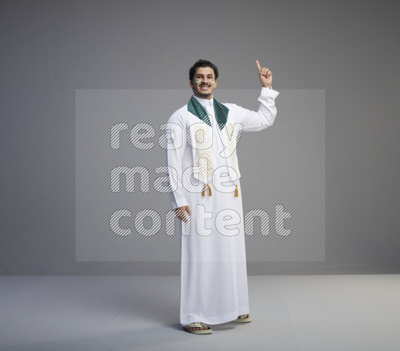 A Saudi man standing wearing thob and Saudi flag scarf with face painting on gray background