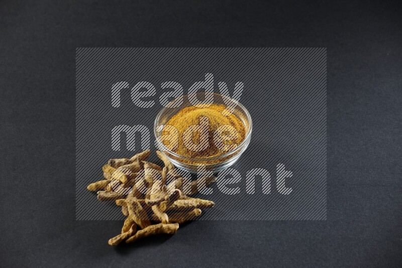 A glass bowl full of turmeric powder and dried whole fingers beside it on black flooring