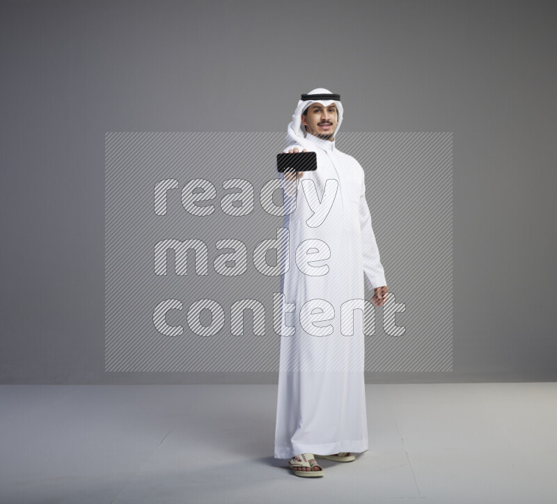 A Saudi man standing wearing thob and white shomag showing phone to camera on gray background
