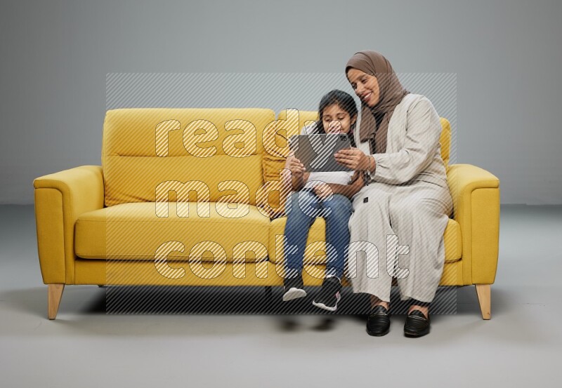 A girl with her mother on yellow sofa and watching on iPad on gray background