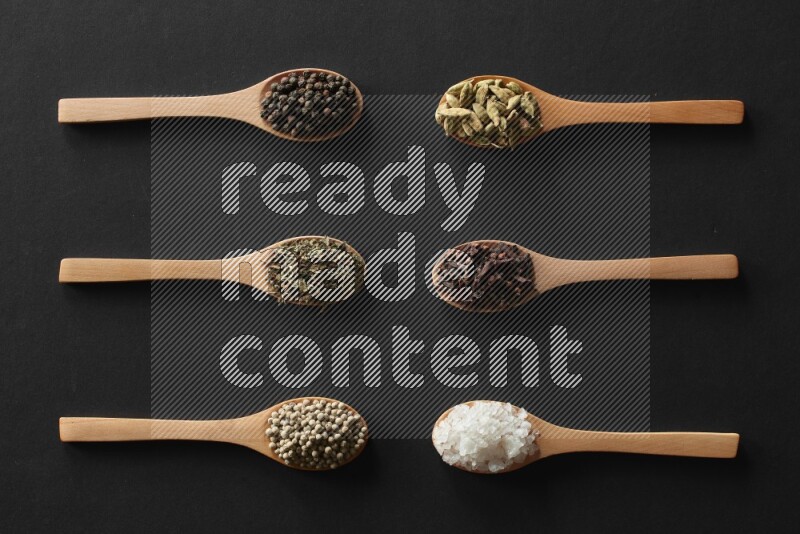 wooden spoons filled with white peppers, cloves, cardamom, salt, black peppers and basil on black flooring