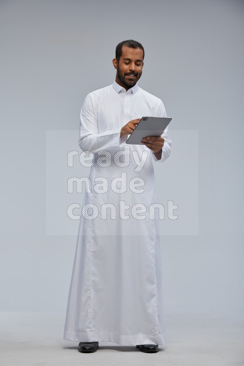 Saudi man wearing Thob standing working on tablet on Gray background
