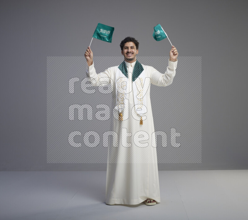 A Saudi man standing wearing thob and saudi flag scarf and holding small saudi flag on gray background