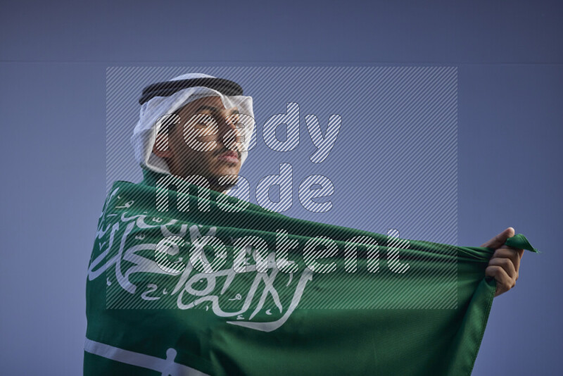 A close-up shot of Saudi man wearing thob and white shomag wrapping big Saudi flag on gray background