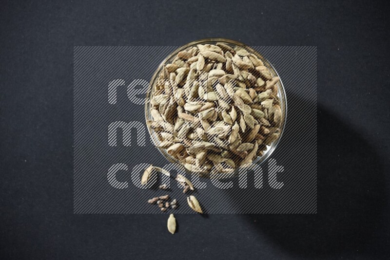 A glass bowl full of cardamom and more seeds spreaded beneath the bowl on black flooring