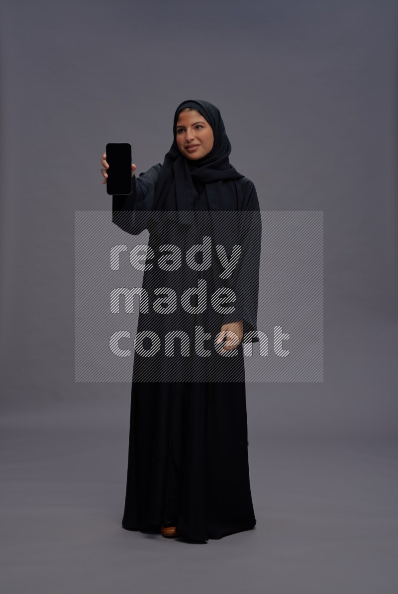 Saudi woman wearing Abaya standing showing phone to camera on gray background