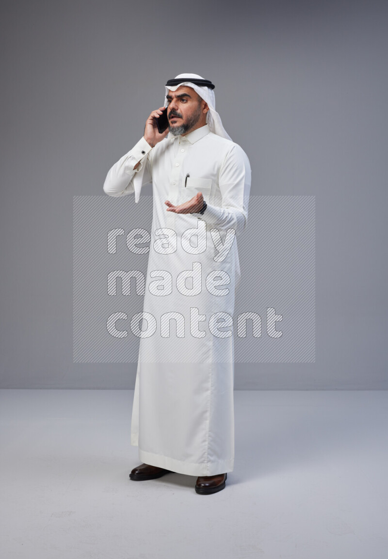 Saudi man Wearing Thob and white Shomag standing talking on phone on Gray background