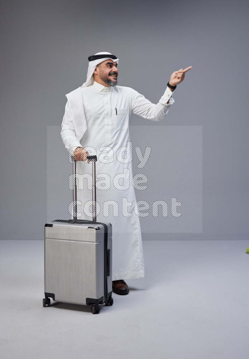 Saudi man wearing Thob and white Shomag standing holding Travel bag on Gray background