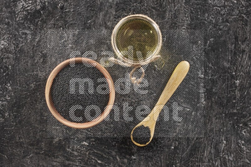 وعاء خشبي وملعقة ممتلئان ببذور حبة البركة وجرة زجاجية من زيت حبة البركة على أرضية سوداء