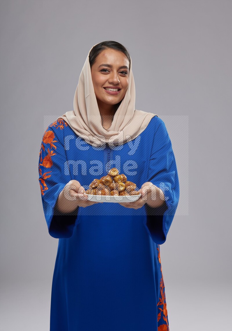 A Saudi woman standing wearing Jalabeya holding a plate of dates