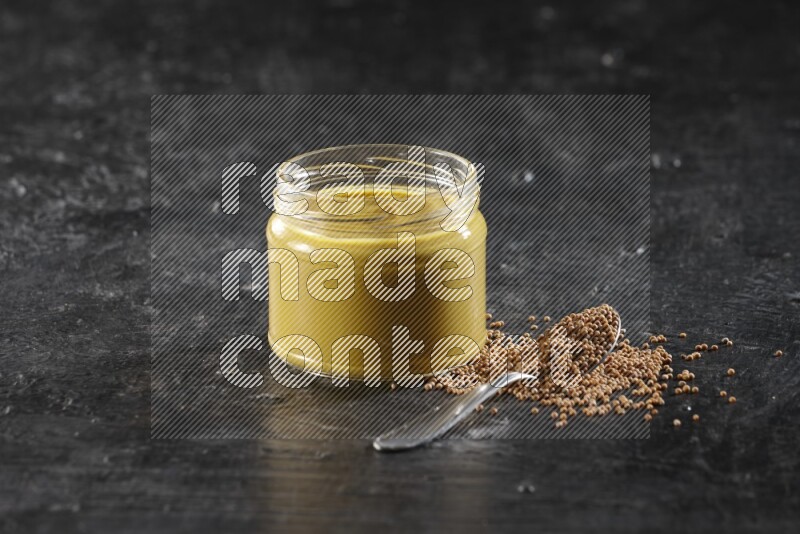 A glass jar full of mustard paste and a metal spoon full of mustard seeds on a textured black flooring