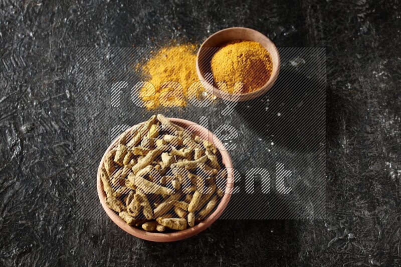 2 wooden bowls, one full of turmeric powder and the other full of dried turmeric whole fingers on a textured black flooring