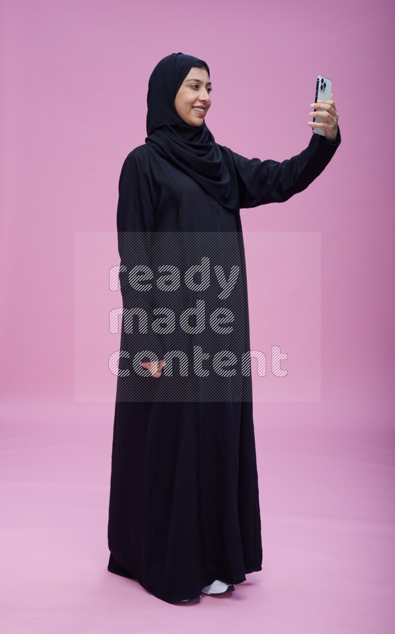 Saudi woman wearing Abaya standing taking selfie on pink background