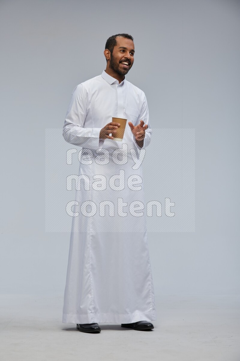 Saudi man wearing thob standing holding paper cup on gray background